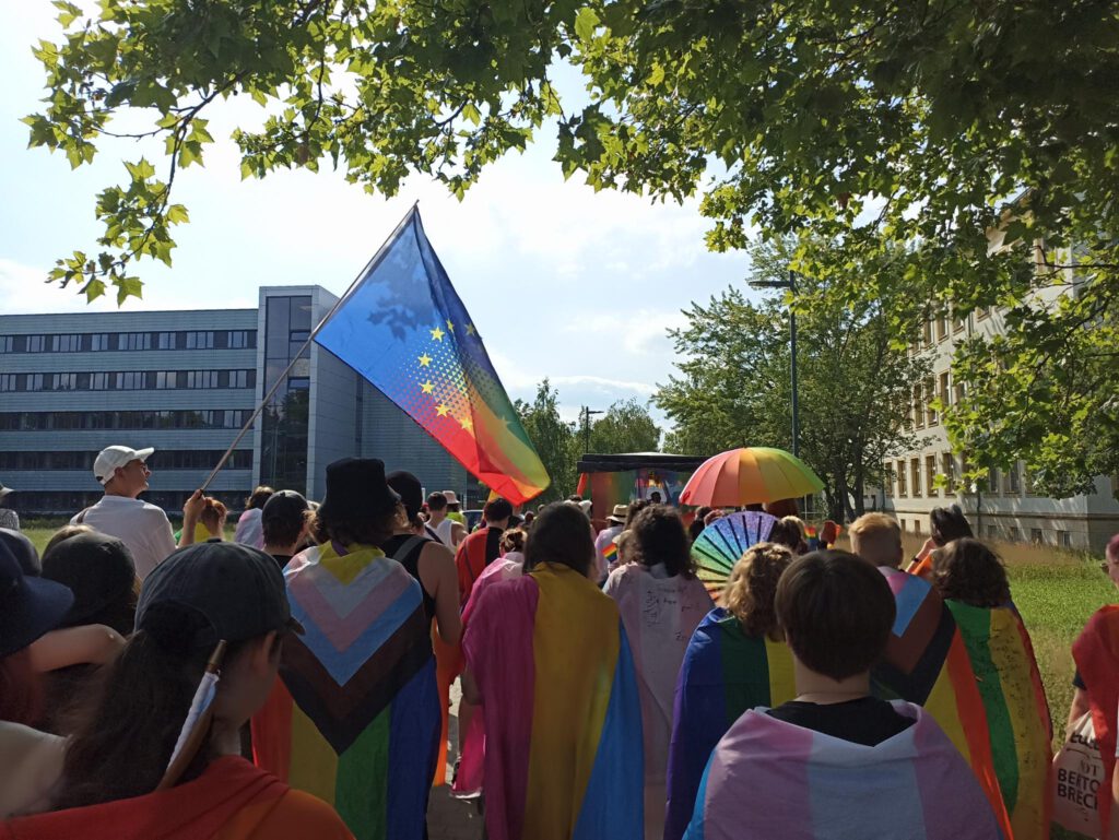 Demozug mit RegenbogenEU Flagge unter Bäumen. Es ist leicht Bewölkt.