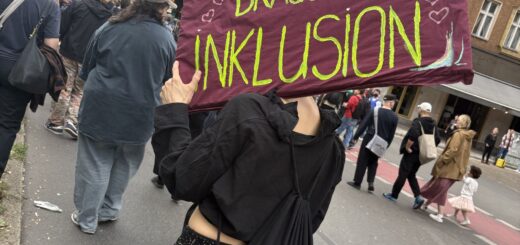 Eine Person in schwarzer Kleidung mit rotem Demoschild. In Neongrün steht darauf: "Demokratie braucht Inklusion"
