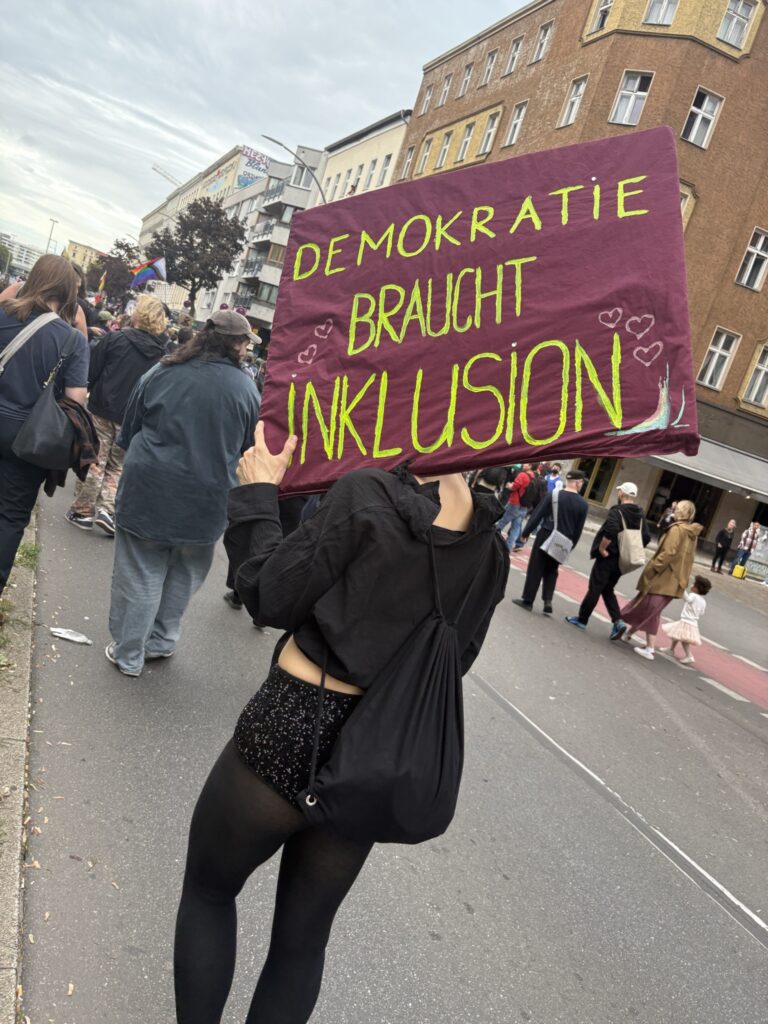 Eine Person in schwarzer Kleidung mit rotem Demoschild. In Neongrün steht darauf: "Demokratie braucht Inklusion"