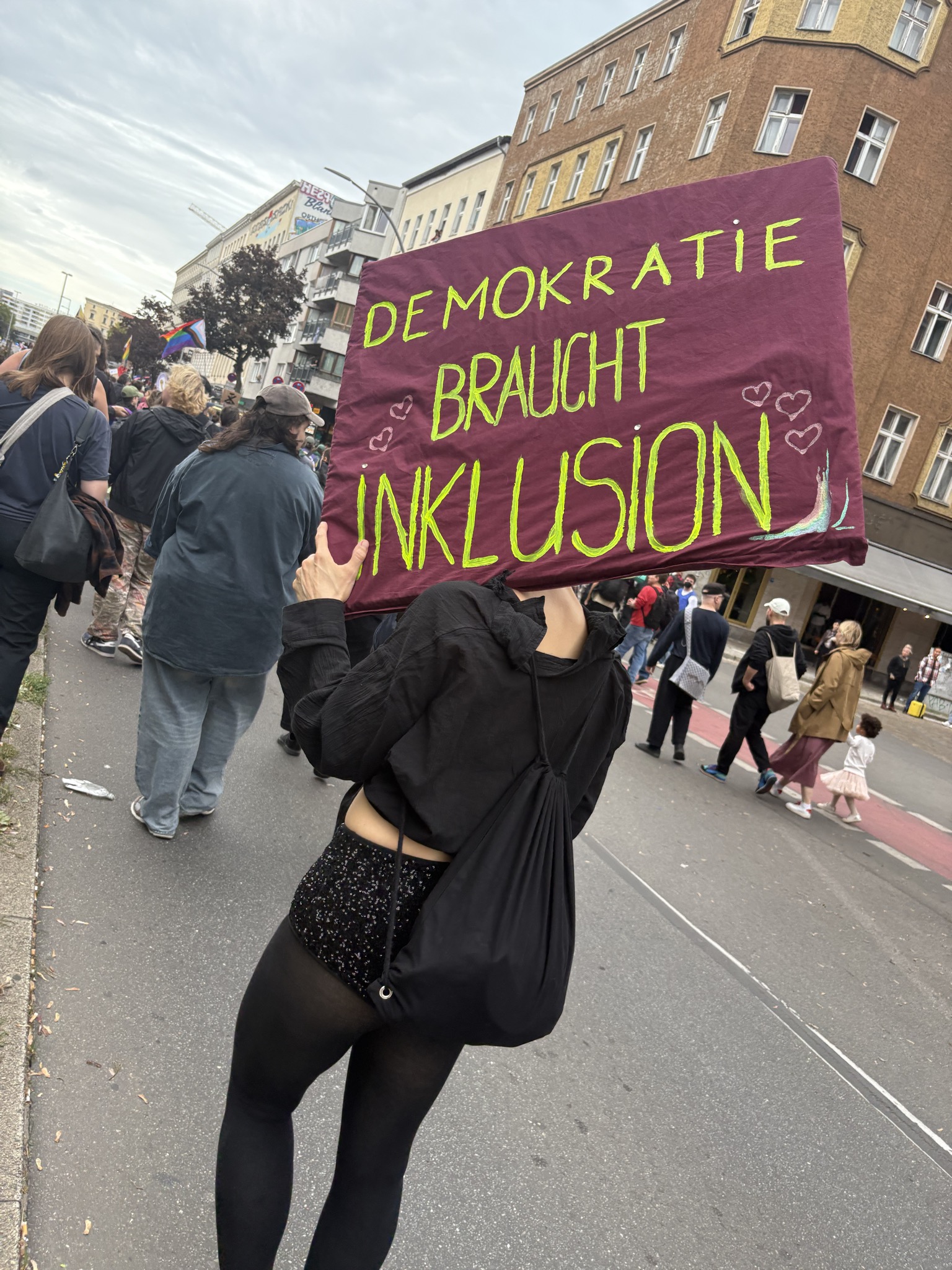 Eine Person in schwarzer Kleidung mit rotem Demoschild. In Neongrün steht darauf: "Demokratie braucht Inklusion"